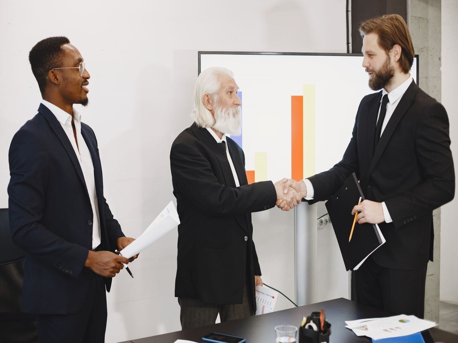escucha activa en negociaciones comerciales, Three businessmen in suits having a meeting, shaking hands in front of a presentation chart.
